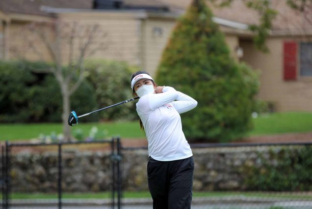 Photo of Zhangcheng Guo (ENG’21) in a visor and white long sleeve, mid golf-swing. Behind her, vegetation and tan building is seen blurred.