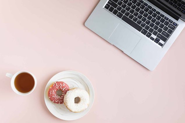 Top view of a cup of coffee, 2 donuts on a plate, and a MacBook laptop sitting on a pink table