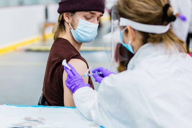 Photo of Ian Forcey, in a brown beanie, looking down at his shoulder, as a medical provider in a face shield, mask and purple gloves gives him a vaccine.