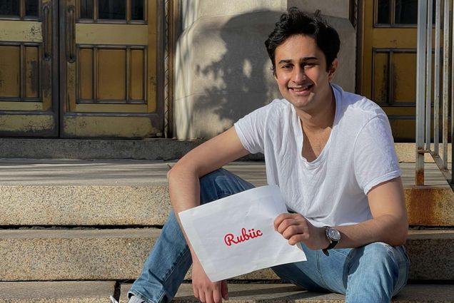 Photo of Asad Malik (CAS’20) sitting on granite steps, with a white t-shirt, sneakers and jeans on, smiling and holding an envelope that says "Rubiic" in red cursive font.