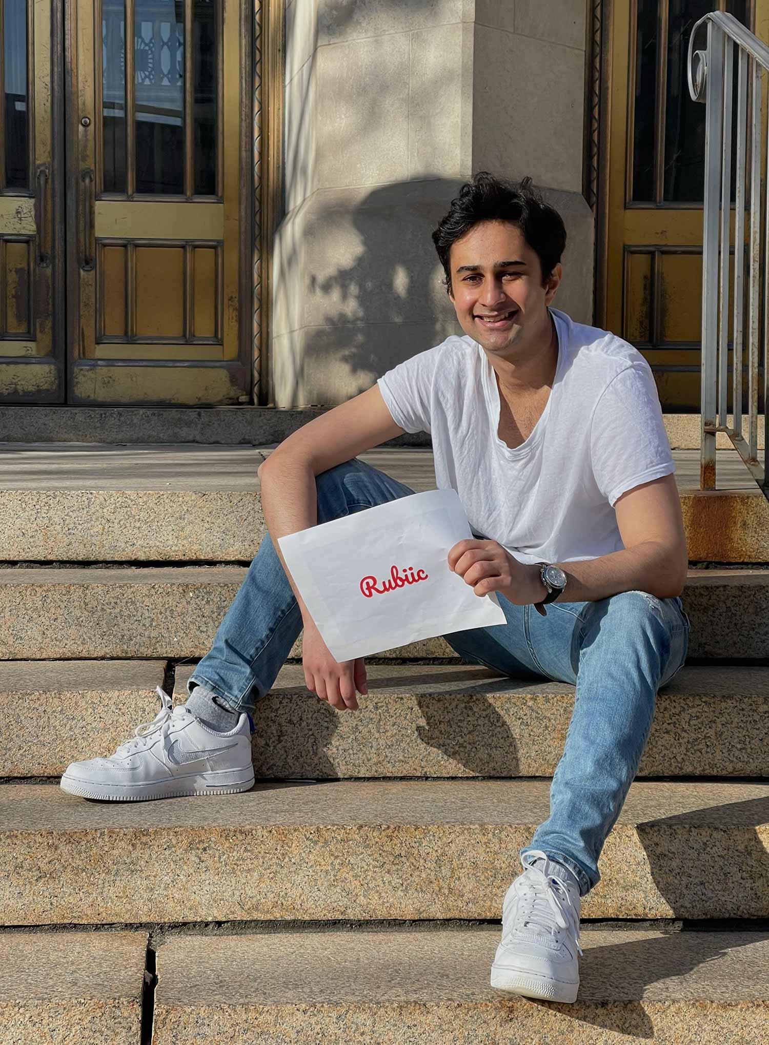 Photo of Asad Malik (MET’21) sitting on granite steps, with a white t-shirt, sneakers and jeans on, smiling and holding an envelope that says "Rubiic" in red cursive font.