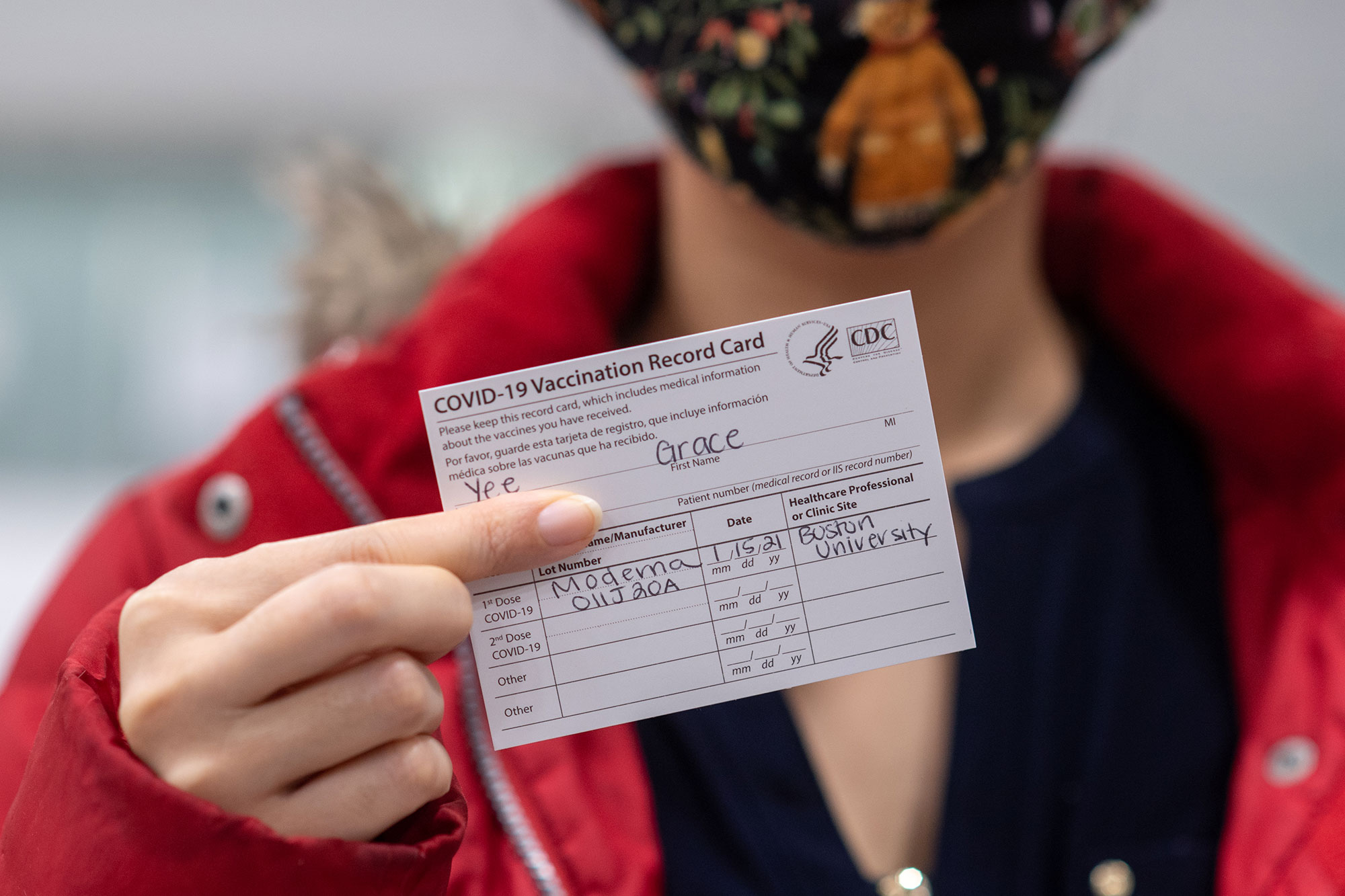 Healthway nurse practitioner case manager Grace Yee does up her CDC Vaccination verification card after receiving the Moderna COVID-19 vaccine January 15 at FitRec. She wears a red winter jacket and a patterned mask. The card is seen in the foreground and she is blurred.