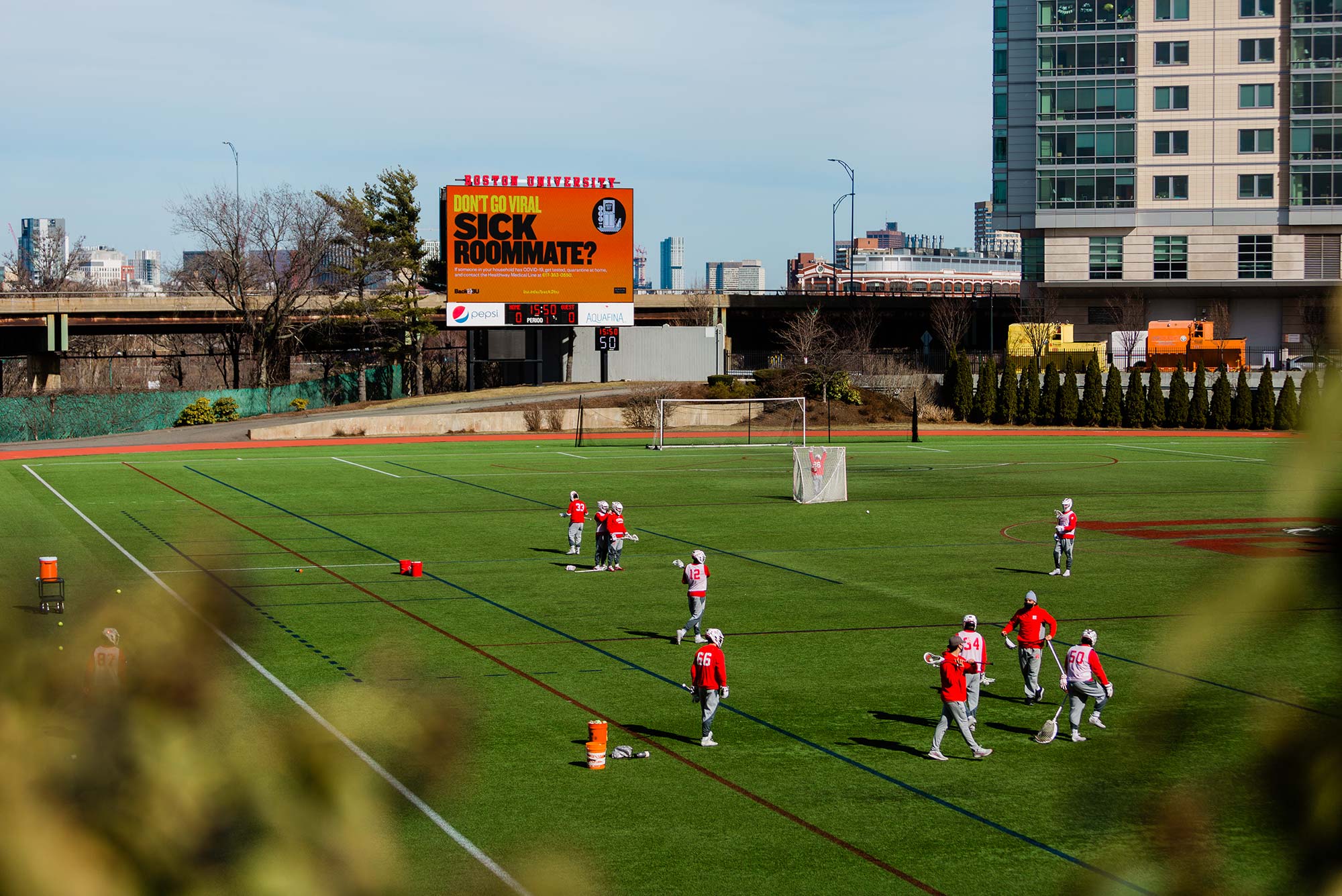 Photo of lacrosse players practicing on Nickerson Field; a large orange billboard in the background says “sick roommate?” The sky is a little cloudy and blue.