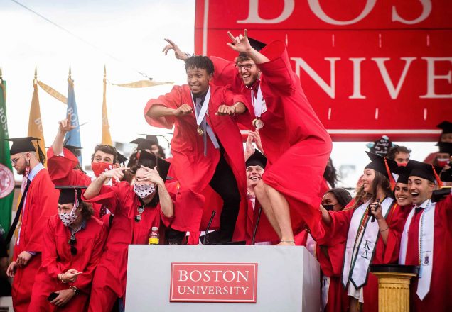 2 students stand on the podium surrounded by a group of graduates posing for a photo after students flooded the stage following Commencement.