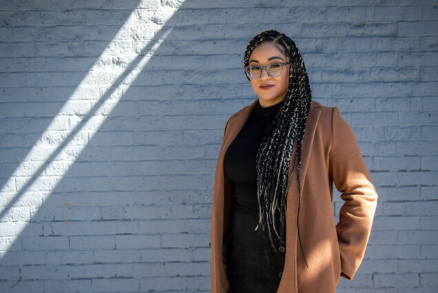 Photo of Ivanna Solano (Wheelock'16) in downtown Lynn. She wears a tan coat and black sweater, and has long braids with patches of gold. She has large glasses and looks earnestly at the camera. Behind her, a ray of sunlight is seen on a brick wall painted light blue.