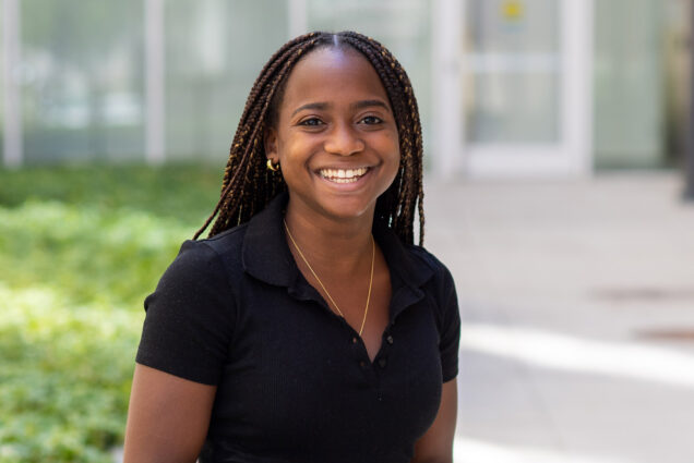 Photo of Archelle Thelemaque (COM’21) in black jeans and a black shirt sitting on a granite bench outside of one of buildings on campus. She has long brown braids and rests her hands on her lap as she smiles.