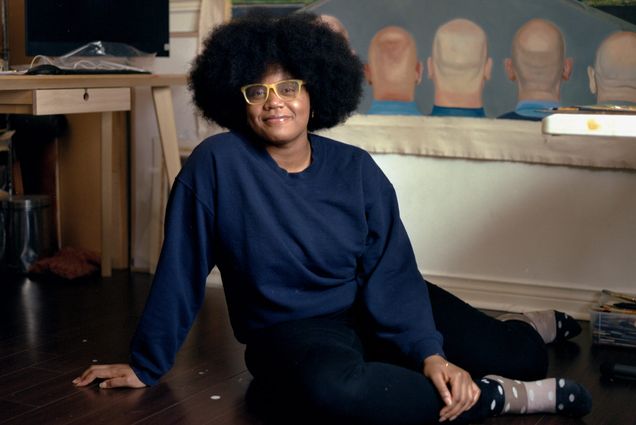 Photo of Shantel Miller (CFA’21) sitting on the ground and smiling. She wears a blue sweatshirt, black pants, polka-dotted socks, and yellow glasses frames. Behind her, a desk with a computer is seen and a painting that shows the back of the heads of a group of white men as two young black women walk down the street.