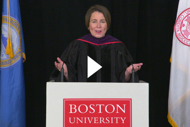 Massachusetts Attorney General Maura Healey during her speech for the LAW convocation. She wears a black robe and stands at a podium with the red Boston University logo on it. To her left is the Boston University flag and to her right, the blue and gold City of Boston flag. Video play button is overlaid.