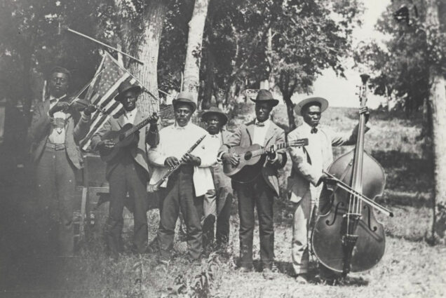 Black and white photograph of a band of African-American playing at Emancipation Day celebration in June 19, 1900, held in “East Woods” on East 24th Street in Austin. There are 6 men in the band, they all wear suits and hats and hold their instruments, including a fiddle, guitar, clarinet, and upright bass. There are trees and an American flag hanging behind them.