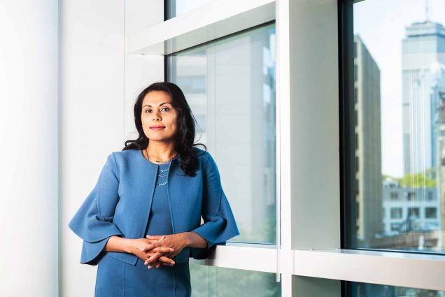 Portrait of Dr. Nahid Bhadelia leaning against a building in front of a window showing a reflection of the Boston skyline.