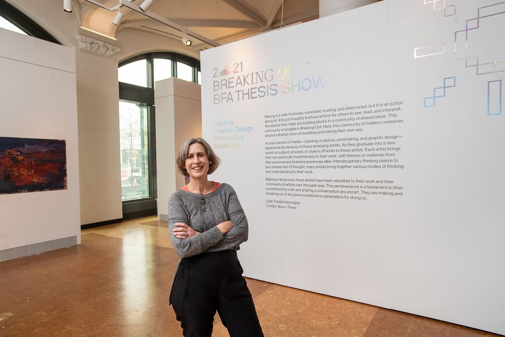 Photo of Dana Clancy, director of the College of Fine Arts’ School of Visual Arts and associate professor of art, in front of the BFA Thesis Exhibition’s curatorial statement in the Stone Gallery. She wears a gray long sleeve blouse and black pants, and smiles with her arms crossed. Behind her, the introductory statement and title of the exhibit, "2021 Breaking Out BFA Thesis Show: Painting, Graphic Design, Printmaking, Sculpture" are seen on the wall.