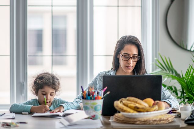 Photo of a young mother sitting a table working on her computer next to a young child who draws in a coloring book.