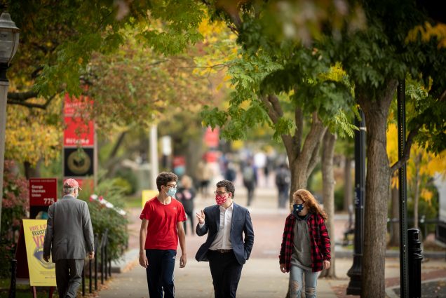 Photo of Isaac Rajagopal (BUA’23) (from left), Chris Kolovos, BU Academy head of school, and Jasper Milstein (BUA’24), strolling down Comm Ave on a fall day. A few people walking are seen walking in the distance. Trees line the Ave and have green and yellow leaves.