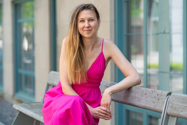 Photo of behavioral scientist Nina Mazar, sitting on a bench, and smiling. She wears a bright pink summery dress and has long blonde hair.