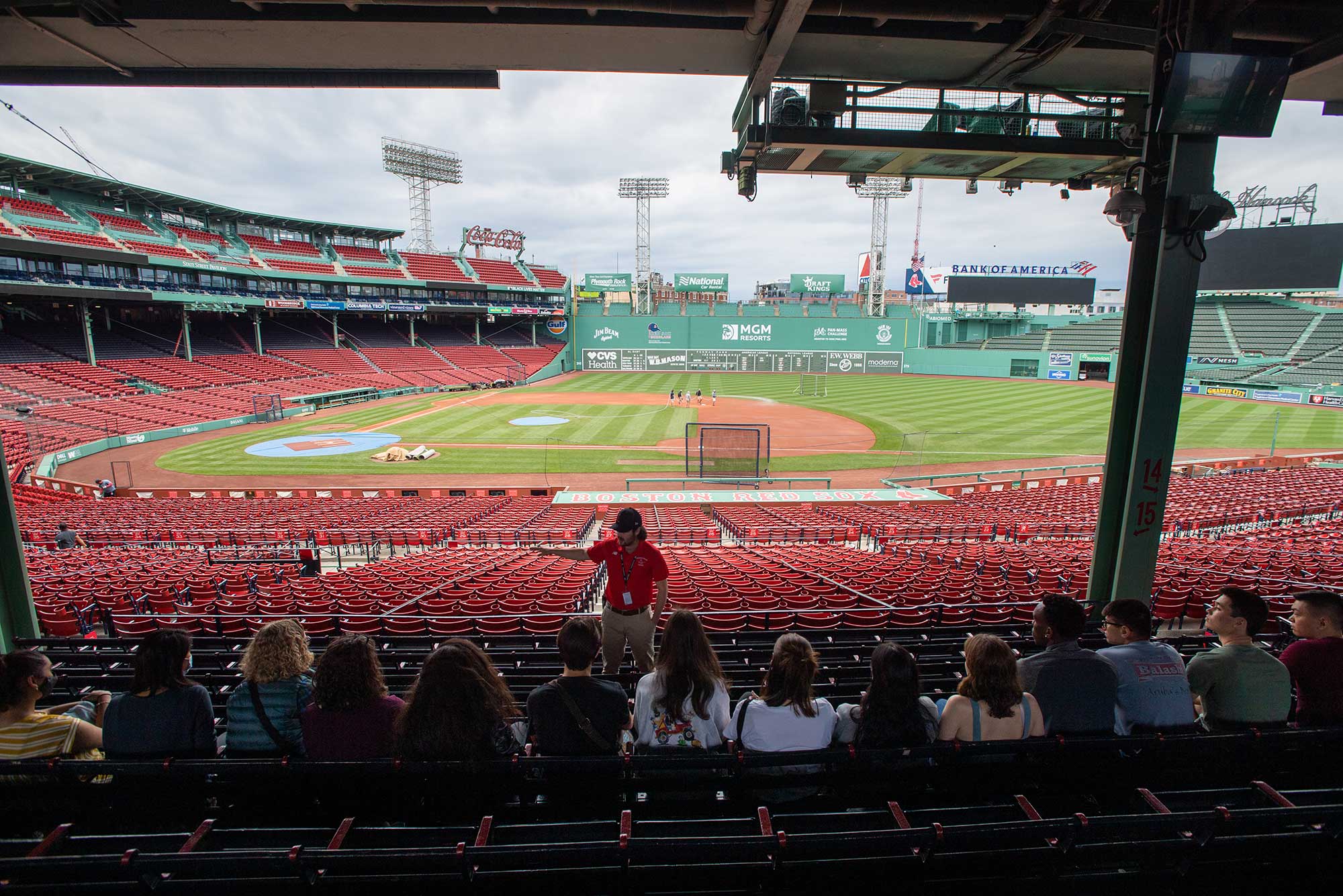 Students sitting in a row of seats at Fenway along the first base line under the overhang as a tour guide dressed in red tells them about the park.