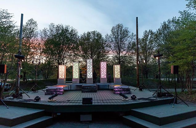 Photo of the stage at the Herter Park Amphitheater. Five illuminated beams ring a small wooden stage; trees and a setting sun are seen in the background.