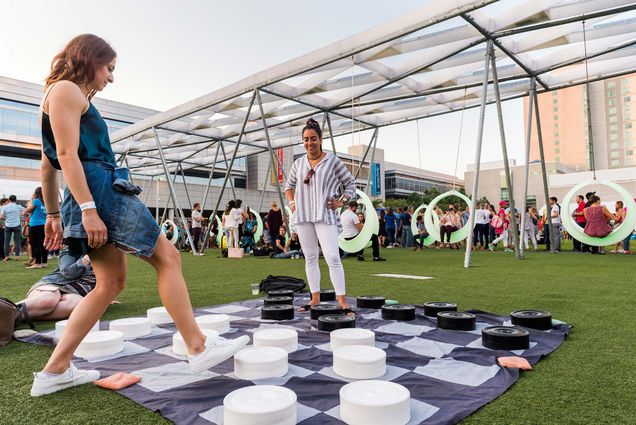 Photo of two women playing outdoor checkers at the Lawn on D. Behind them, people swing on large circular swings.