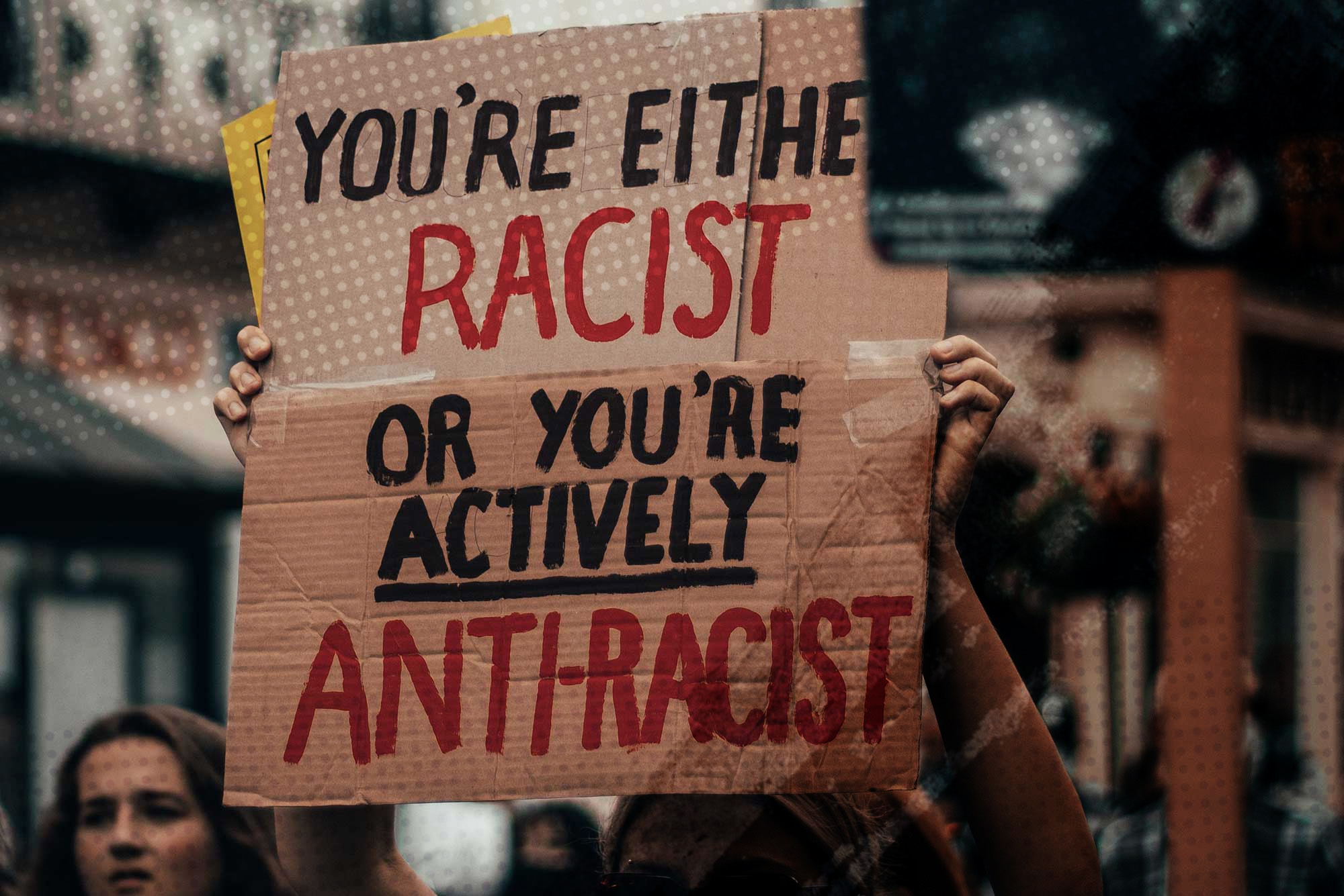 Photo of a woman holding a sign that says "You're either racist or you're actively anti-racist" at a protest.