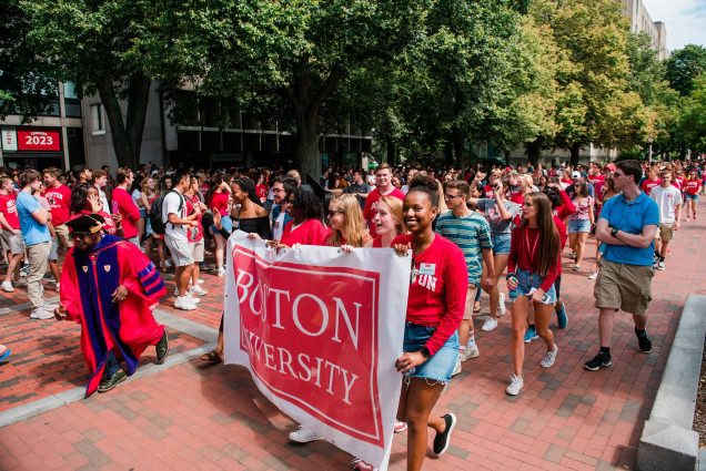 Photo of students carrying a Boston University banner as they walk down Comm Ave. Most students wear red and a faculty in a cape and gown is seen on the left.