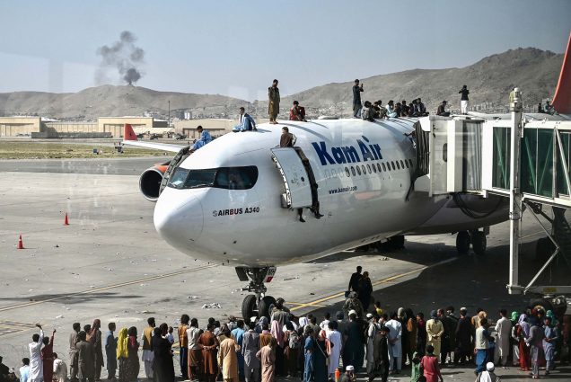 Photo of Afghan people climbing atop a plane, that has a blue 'KamAir' logo on the side, as they wait at the Kabul airport in Kabul on August 16, 2021, as thousands of people mob the city's airport trying to flee the Taliban. People also gather below the plane on the tarmac.