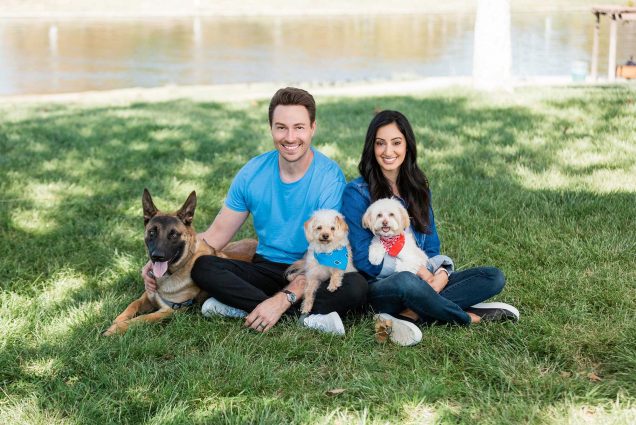 Photo of Rashi Khanna Wiese (CAS’06) and her husband, Eric Wiese, sitting in grass with three dogs, a german shepherd and two smaller dogs that look like they may be part maltese.