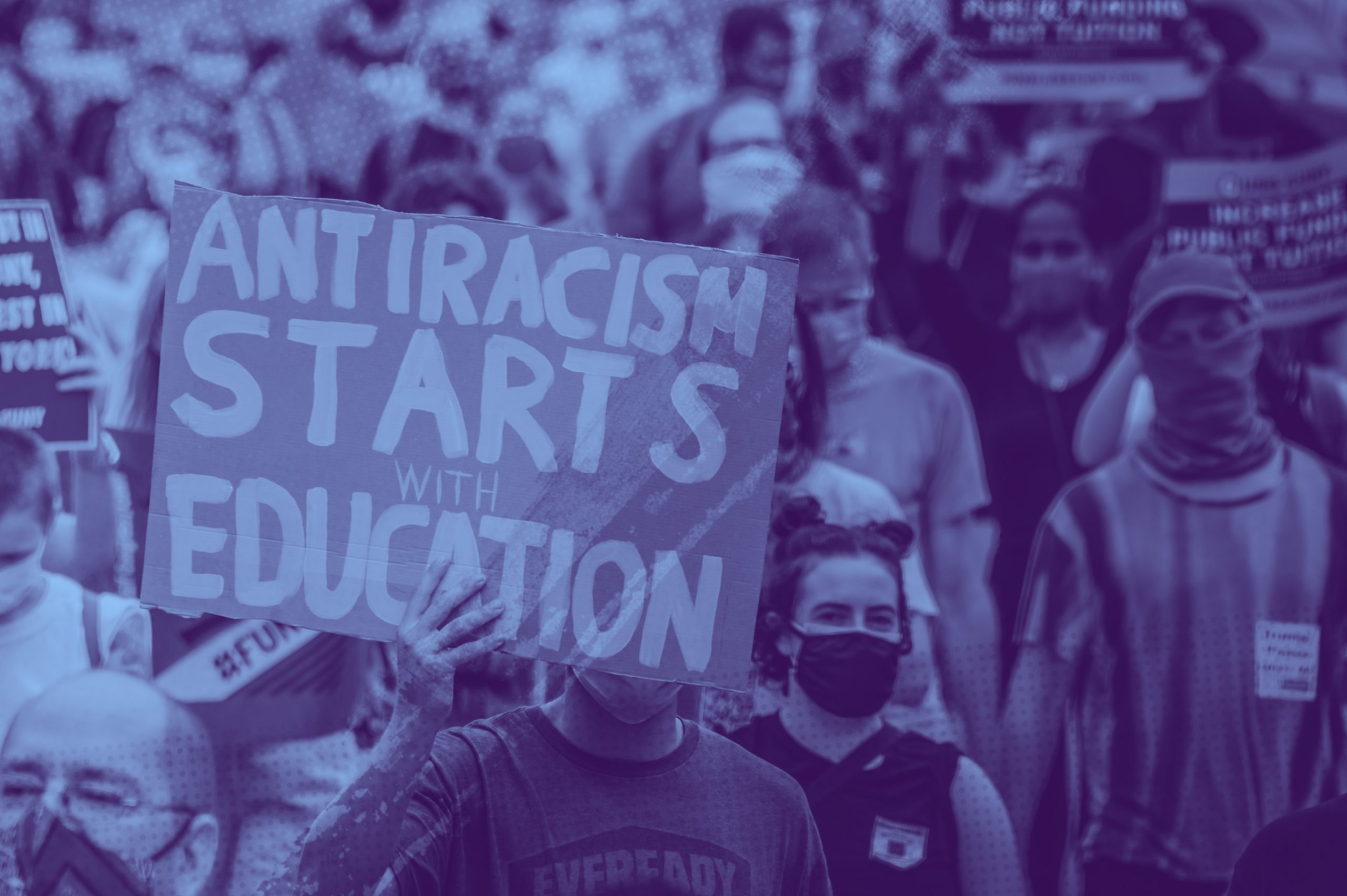 A crowd of people marching at a Black Lives Matter protest. A man holds a sign that says "Antiracism starts with education" at the front of the crowd.