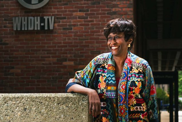 Photo of Boston journalist and COM alumn Carmen Fields posing outside of WHDH in Boston on June 3, 2021. She wears a colorful sweater with a variety of geometric and organic patterns and leans on a cement wall and smiles.