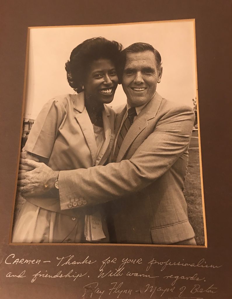 Photo of Boston Mayor Ray Flynn with TV news anchor Carmen Fields in the late 1980s. Flynn gives Fields a side hug and both a dress in suits. The photo has a brown frame and is signed at the bottom by Ray Flynn, it reads in cursive writing in white: "Carmen, thanks for your professionalism and friendship. With warm regards, Ray Flynn, Mayor of Boston." 