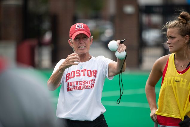 Photo of Sally Starr, wearing a red BU hat and a white Boston University t-shirt with red writing, holding two field hockey balls and a coach's whistle in her hand during what appears to be a field hockey practice.