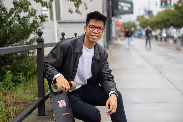 a photo of smiling student vlogger Kreiver Zhou sitting on bench and holding his skateboard during a quiet time on Comm Ave.