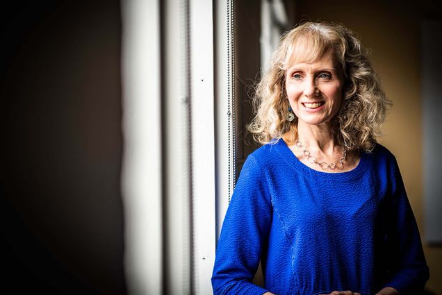 Photo of Deborah Carr, a white woman with wavy blonde hair, smiling and posing for a photo next to a window.