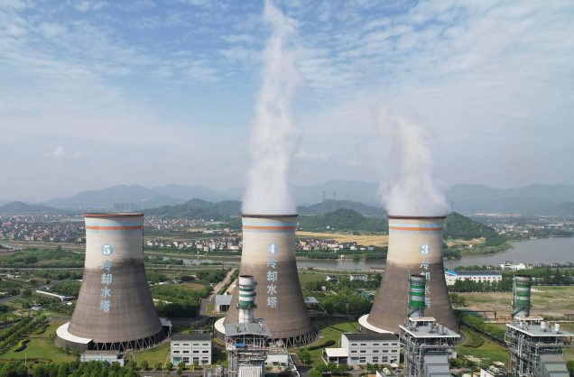 a photo of steam billowing out of grey chimney's of a coal-fired power plant in China. The chimneys feature light blu writing and contrast against a blue, lightly clouded sky.