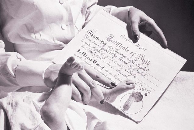 photo of a nurse IMPRINTING a BABYS FOOT ON a birth CERTIFICATE. The document is handwritten, but has "Certificate of Birth" printed in a cursive font at the top. The nurse has painted fingernails.