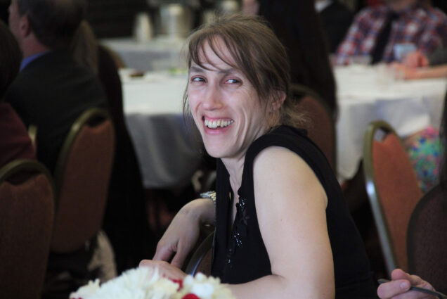 Photo of Jennifer Formichelli, smiling, at a formal dinner. She wears what looks like a black dress and sits at a table; other tables with white table cloths are seen behind her.