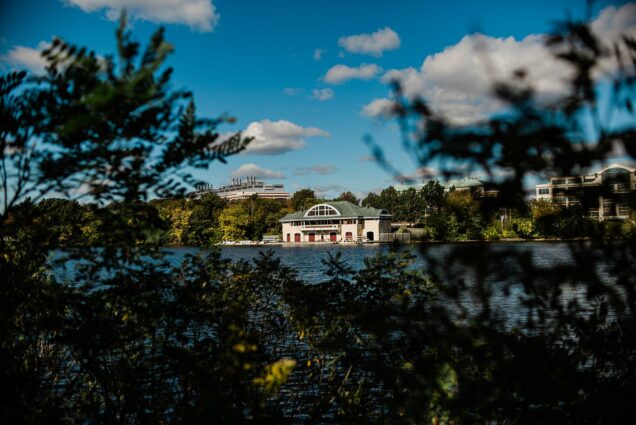 Photo of DeWolfe Boathouse taken from across the Charles River on a sunny fall day. Small clouds are seen in a brilliant, blue sky. The Boathouse is tan with a greenish gray roof.