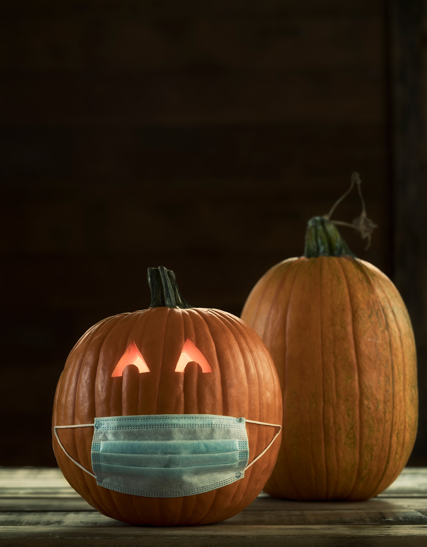 Photo of a Jack O' Lantern with Protective Face Mask with its eyes lit up. A taller pumpkin rests behind it to the right. The background is black.