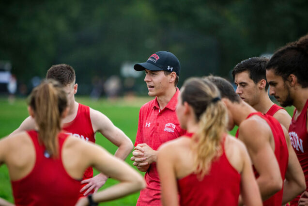 Photo of Jordan Carpenter, new associate head coach of cross country, with a blue Boston University hat on, speaking with cross country runners, dressed in red running uniforms.