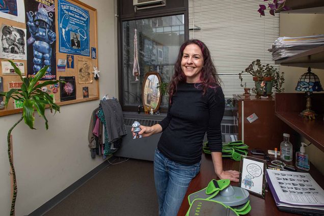 Photo of Creative writing prof Joelle Renstrom in her CGS office for Office Artifacts. She has brown, wavy shoulder length, hair with streaks of purple, and wears a black long-sleeve shirt and jeans, as she holds a small toy robot in her hand.