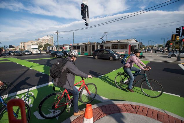 Photo of bikers using the bike lanes at the intersection of Commonwealth Ave and Montfort Street. Both men wear helmets as they use the lanes. In the background, cars drive and the green line T go past.