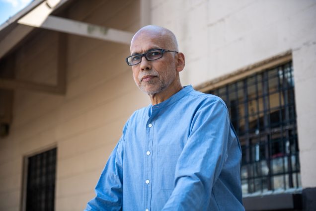 photo of André de Quadros, choir director and teacher of music in the BU prison education program, looking at the camera with a stoic expression. He wears black, rectangular glasses and a light blue collared shirt.