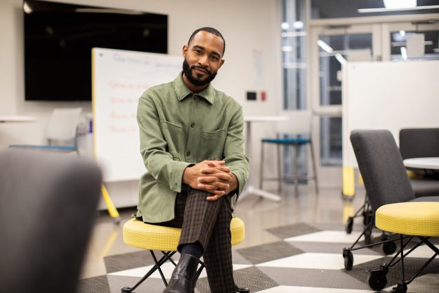 Photo of Jonathan Allen (LAW’19), a young Black man with a beard, sitting on a yellow stool. He wears a green jacket and smiles. A whiteboard is seen behind him.