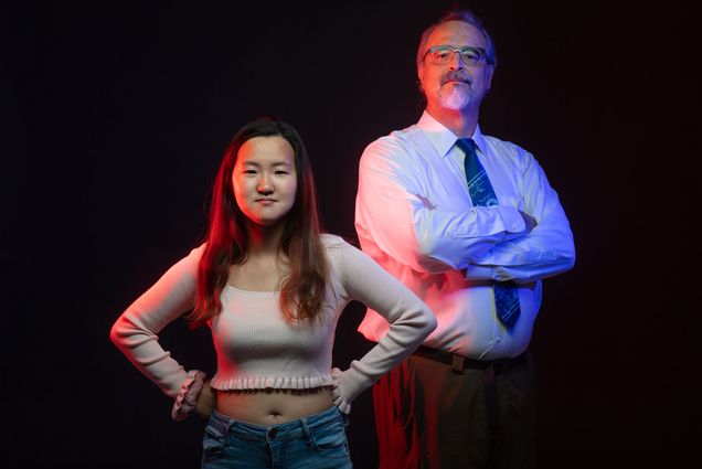 Studio photo of CGS senior lecturer Charles Henebry and student Rachel Do (CGS’22). Rachel stands in front in a Wonder Woman pose and Henebry stands slightly behind her and to the right with arms crossed and head tilted slightly up. Both where stoic faces as green and blue lights cast a heroic light on them.