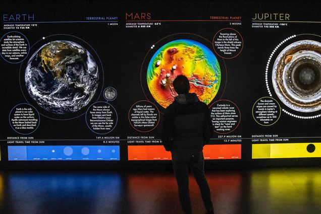 A visitor views a Green Zone exhibit during the COP26 climate talks in Glasgow, U.K., on Monday, Nov. 1, 2021. He stands in a dark room facing a bright, colorful display showing the differences in climate on Earth, Mars, and Jupiter.