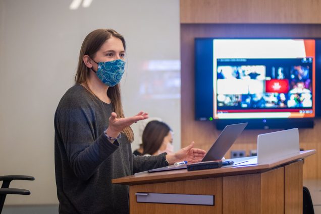 Photo of Lecturer Malwina “Maja” Carrion in her course Neglected Tropical Diseases at Sargent Nov 9. She wears a patterned, multi-color, bluish mask, and a gray long sleeve, and gestures with her right hand at the podium at the front of the classroom. She is a young light-skinned woman with long hair.