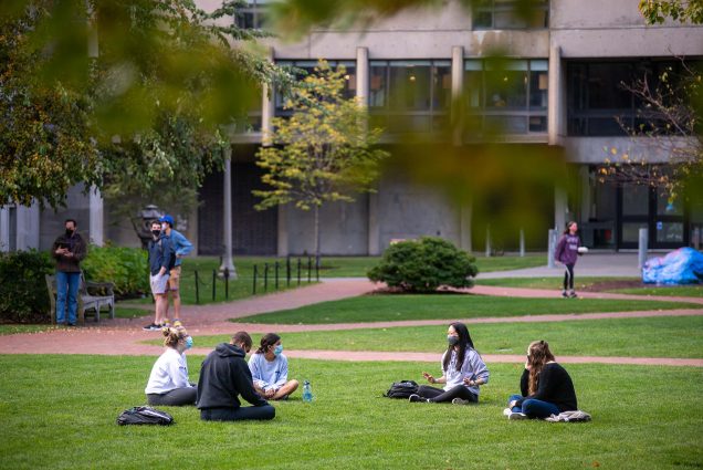 Photo of groups on BU Beach on Oct 23, 2020 safely conversing. A group of five seated in the grass sits socially distanced. In the background other students are seen conversing in small groups with masks on.