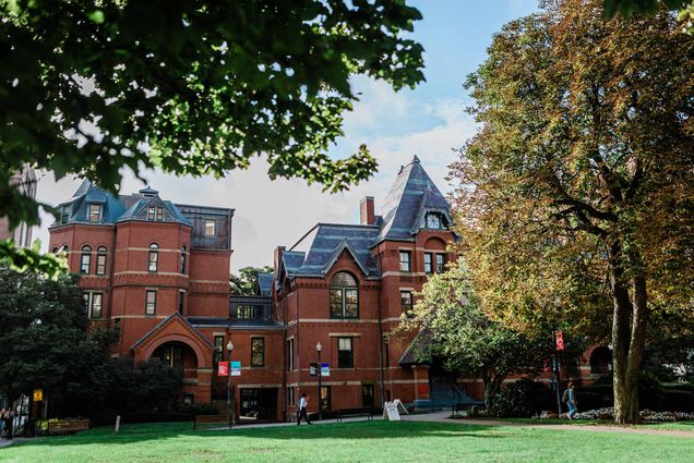 photo of the Boston University School of Public Health, an early 20th century brick building with a small gothic pattern and gray/blue roof shingles. The grass out front is bright green, and a student walks along a path near the building.