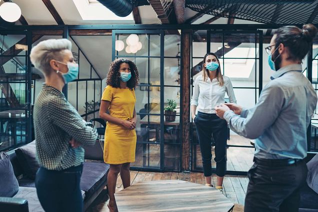 stock photo of a diverse group of business people with protective masks meeting in a co-working area. They are standing socially distanced and all wear business casual attire.