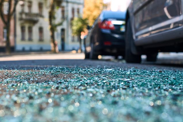 photo of a car crash. Angle is low, showing shattered glass on the road as a blurry background shows a fender bender during the day.