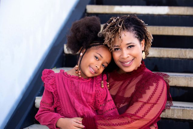 Photo of Zenda Walker in a red blouse, smiling, as she sits on a staircase with her daughter, in pink, in her lap.