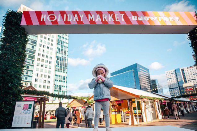 Photo a small child drinking a sippy cup underneath a sign that reads "Holiday Market: Snowport" with pink and red stripes.